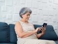 Happy Asian senior woman sit on couch, holding and looking at digital tablet screen in hand in living room. Royalty Free Stock Photo