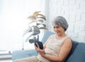 Happy Asian senior woman sit on couch, holding and looking at digital tablet screen in hand in living room. Royalty Free Stock Photo