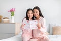 Happy asian teen girl and young woman reading postcard sitting on bed in minimalist bedroom interior