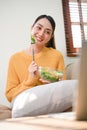 Happy Asian woman eating salad bowl while enjoys watching movie via laptop on sofa Royalty Free Stock Photo