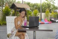 Happy Asian woman in elegant and dress sitting outdoors at pool resort coffee shop having healthy fruit juice working on lapt Royalty Free Stock Photo