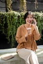 A happy Asian woman is enjoying talking on the phone and sipping coffee while relaxing on a bench Royalty Free Stock Photo