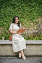 A happy Asian woman enjoys reading a book on a bench in a city park on her weekend