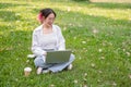 A happy Asian woman in glasses and casual wear sits on the grass in a park working on her laptop Royalty Free Stock Photo