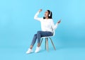 Happy Asian woman sitting on chair wearing wireless headphone listening to music from smartphone on blue background Royalty Free Stock Photo