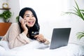 Happy Asian woman talking on mobile phone shopping online with credit card lying down in front of laptop in bed Royalty Free Stock Photo