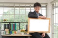 Happy Asian young businessman holding an empty white board and sitting on the table in office. Concept of copy space for advertise Royalty Free Stock Photo