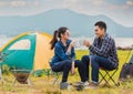 Happy Asian young couple sitting on picnic chair drinking tea and coffee while tent camp lakeside at parks outdoors on vacation