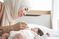 Happy Asian young Muslim mother wear hijab holding milk bottle, feeding milk her cute newborn baby 1 month old, lying on bed at Royalty Free Stock Photo