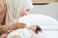 Happy Asian young Muslim mother wear hijab holding milk bottle, feeding milk her cute newborn baby 1 month old, lying on bed at Royalty Free Stock Photo