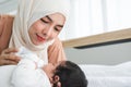 Happy Asian young Muslim mother wear hijab holding milk bottle, feeding milk her cute newborn baby 1 month old, lying on bed at Royalty Free Stock Photo