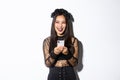 Happy attractive asian woman in halloween costume laughing carefree, holding smartphone and looking amused at upper left