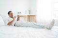 A happy attractive bearded Caucasian guy laying on the bed with vape in apartment in the morning, using a smartphone.