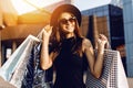 Happy attractive elegant girl, in a black dress and hat, wearing dark glasses, holding shopping bags and enjoying shopping. Black Royalty Free Stock Photo