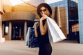 Happy attractive elegant girl, in a black dress and hat, wearing dark glasses, holding shopping bags and enjoying shopping. Black Royalty Free Stock Photo