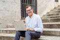 Happy attractive mature man working on laptop checking email sitting on stairs outdoors urban area Royalty Free Stock Photo