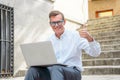 Happy attractive mature man working on laptop checking email sitting on stairs outdoors urban area Royalty Free Stock Photo