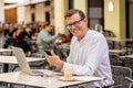 Happy attractive stylish mature man sending message on mobile while working on laptop outside cafe Royalty Free Stock Photo