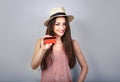 Happy attractive woman in summer hat holding and showing red credit card on grey background Royalty Free Stock Photo
