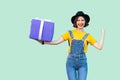 Happy attractive young girl in hipster wear in denim overalls and black hat standing and holding big heavy gift box with toothy