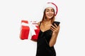 Happy attractive young woman in red christmas hat holding gift box and using mobile phone isolated on white background Royalty Free Stock Photo