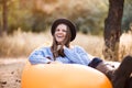 Happy autumn portrait of young woman in blue sweater and black hat lying on orange inflatable sofa outdoor in forest Royalty Free Stock Photo