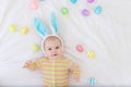 Happy baby boy with rabbit ears on his head lying on the bed with Easter eggs, cute funny smiling little baby. The concept of Royalty Free Stock Photo