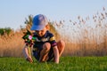 Happy baby boy sitting in grass on the fieald at sunny summer evening. Child outdoors with wind wheel on the river bank