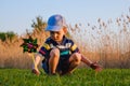 Happy baby boy sitting in grass on the fieald at sunny summer evening. Child outdoors with wind wheel on the river bank