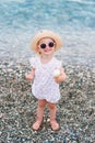 Happy baby girl dressed in summer clothes, yellow hat and pink sunglasses stands on the beach eats white ice cream and Royalty Free Stock Photo