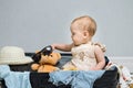 Happy baby girl sitting in travel suitcase puts sunglasses on a bear toy. Child with amused expression enjoys