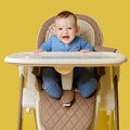 Happy baby sits on a high chair for feeding children, studio yellow background. Smiling child boy at the age of six months eating Royalty Free Stock Photo