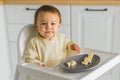 Happy baby sitting in high chair eating fruit in kitchen. Healthy nutrition for kids. Bio carrot as first solid food for Royalty Free Stock Photo