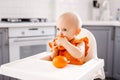 Happy baby sitting in high chair eating fruits in a white kitchen. Healthy nutrition for kids. Children eat oranges and Royalty Free Stock Photo