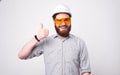 A happy bearded architect wearing a helmet and a pair of protection glasses is showing a thumb up to the camera near a Royalty Free Stock Photo
