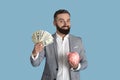 Happy bearded entrepreneur in formalwear holding cash and piggybank against blue studio background Royalty Free Stock Photo