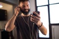 Happy bearded guy wearing casual grey t-shirt listening music in earphones, checking social networks on smartphone Royalty Free Stock Photo