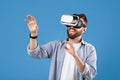 Happy bearded young man using VR headset to explore augmented reality on blue studio background Royalty Free Stock Photo