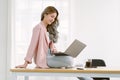 Happy beautiful Asian casual freelancer sitting on top desk using laptop computer in bright room at home office. Young stylish Royalty Free Stock Photo