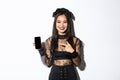 Happy beautiful asian girl in witch costume pointing finger at smartphone screen with pleased smile, showing halloween