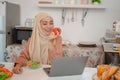 A happy Asian Muslim woman in a hijab is working on her laptop at a dining table in the kitchen Royalty Free Stock Photo