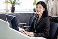 Happy beautiful Asian woman officer typing on desktop computer while sitting at office desk with blurred background of her busy Royalty Free Stock Photo