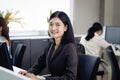 Happy beautiful Asian woman officer typing on desktop computer while sitting at office desk with blurred background of her busy Royalty Free Stock Photo