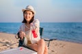 Happy beautiful Asian woman sitting on picnic chair wear sunglasses and hat holding red beverage bottle on beach in outdoor