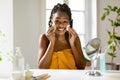 Happy beautiful black lady sitting in front of mirror in bedroom, erasing make up from her face, using cotton pads Royalty Free Stock Photo