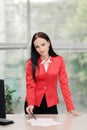 A young attractive Caucasian blond woman in a red business suit sits at a desk in a bright office. Portrait of a Royalty Free Stock Photo