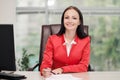 A young attractive Caucasian blond woman in a red business suit sits at a desk in a bright office. Portrait of a Royalty Free Stock Photo