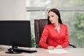 A young attractive Caucasian blond woman in a red business suit sits at a desk in a bright office. Portrait of a Royalty Free Stock Photo