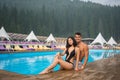 Happy beautiful couple sitting on the edge of swimming pool in the luxurious resort. On the background of mighty forests Royalty Free Stock Photo