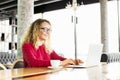 Happy beautiful curly hair blonde young female at coffee shop using laptop, smiling. Young attractive woman freelancer wearing gla Royalty Free Stock Photo